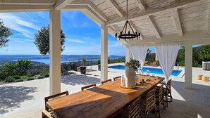 Un espace extérieur abrité, aménagé pour les repas, avec une table et des chaises en bois, offrant une vue sur une piscine et un paysage pittoresque de la mer et des collines.