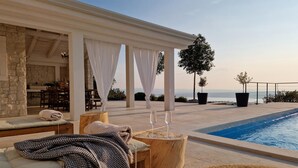 Un espace aménagé au bord de la piscine, avec un coin salon couvert, une table en bois et deux verres, et une vue sur la mer.