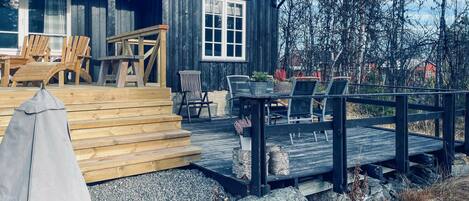 Una terraza de madera con mobiliario exterior, una zona de estar cubierta y vistas a un área boscosa.