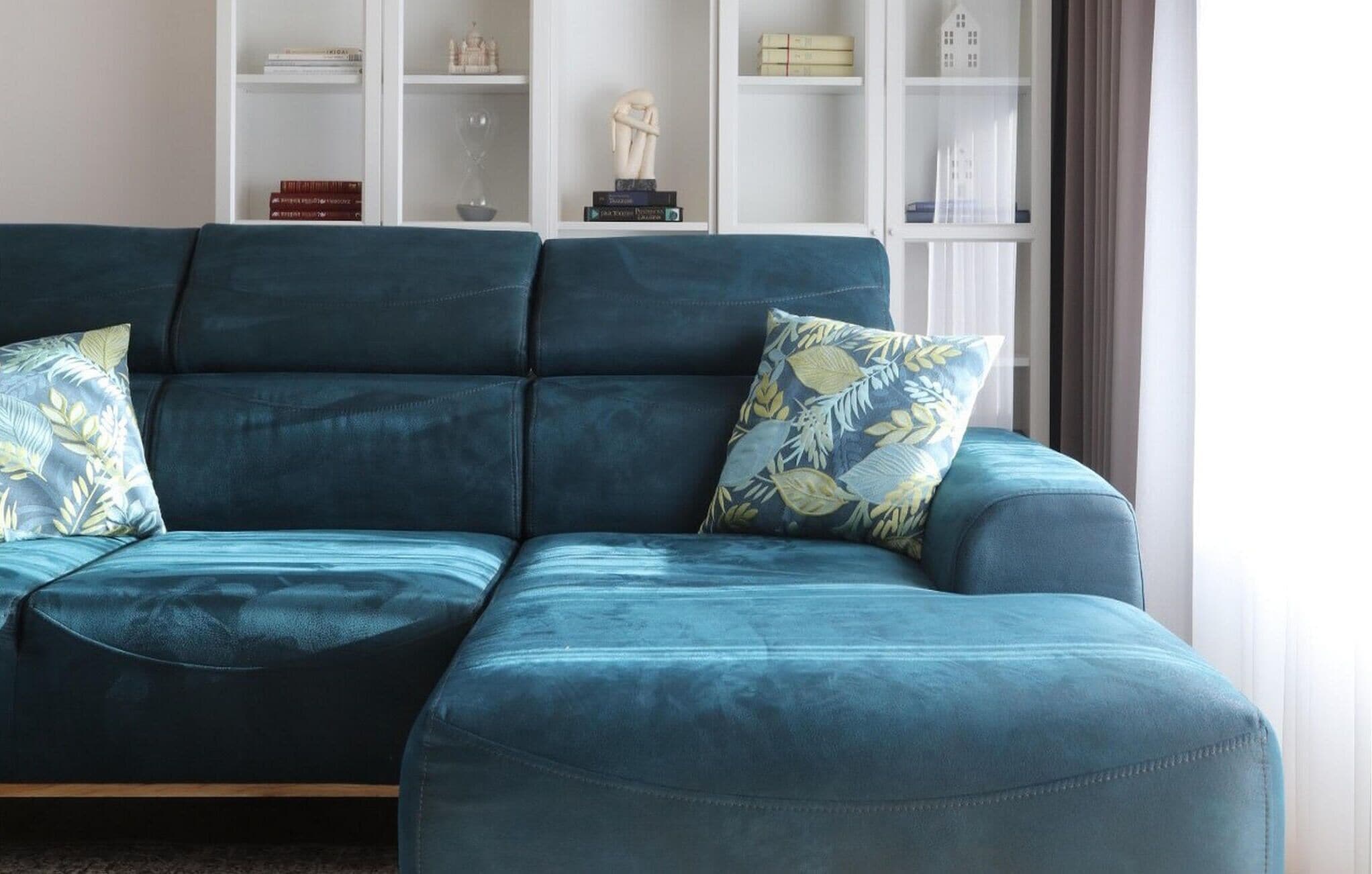 Un divano modulare color petrolio con cuscini a motivi floreali, una scaffalatura bianca con oggetti decorativi e una finestra con tende.