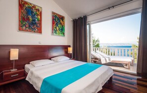 A hotel room with a bed, a nightstand, a lamp, a view of the sea, and two abstract paintings on the wall.