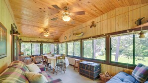 A cabin interior with a dining area, two sofas, a ceiling fan, and large windows. - Grill & Fire Pit: Waterfront Cameron Retreat! (Cameron)