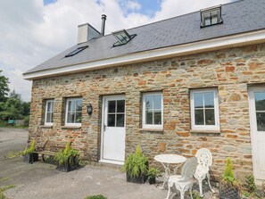 A stone building with two windows, a door, and a small outdoor seating area with a table and chairs.
