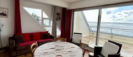 Een kamer met een ronde tafel, stoelen, een bank en grote ramen met uitzicht op zee.