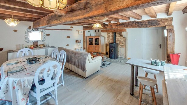 A rustic interior with exposed wooden beams, a dining area with white chairs and a patterned tablecloth, a sofa, and a small kitchen area with a wood-burning stove.