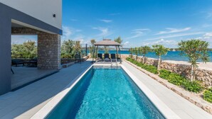 Una piscina con un pergolato e poltrone relax, circondata da mura in pietra e vegetazione, con vista sul mare.