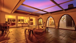 A spacious indoor area with a high ceiling, a skylight, and a view of the sky. There are multiple seating areas with tables and chairs, and a decorative fountain. - 5BR/4BA |Pool & Spa + Pickleball, Golf View (Sarasota)