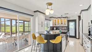 A modern kitchen with a central island, bar stools, and a view of a golf course through large sliding doors. - 5BR/4BA |Pool & Spa + Pickleball, Golf View (Sarasota)