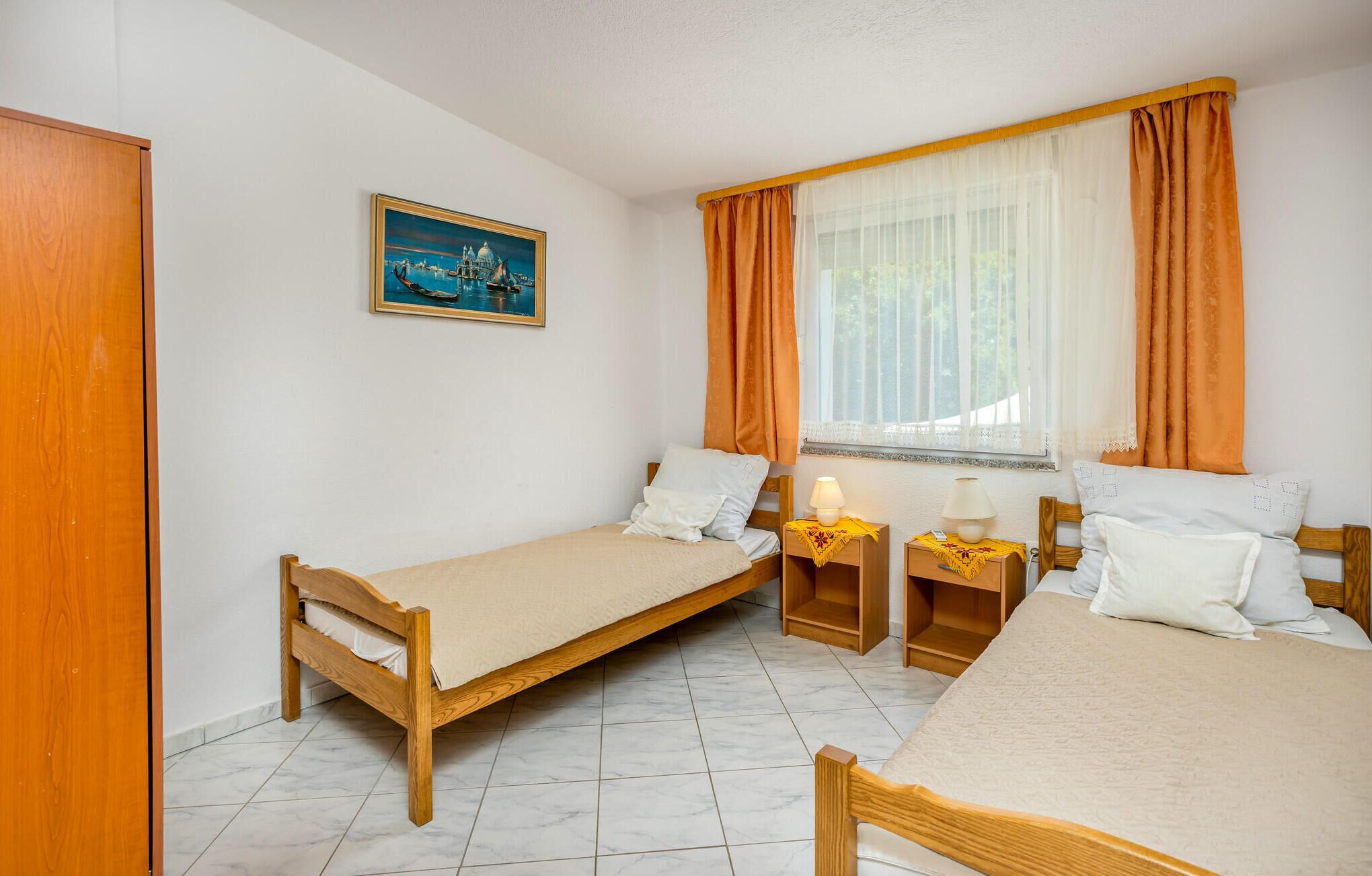 Ein Zimmer mit zwei Holzbett, einem Fenster mit orangefarbenen Vorhängen, einem Nachttisch mit Lampe und einem Bild an der Wand.