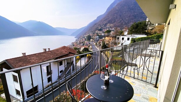 Un balcón con mesa y sillas, con vista a un lago y un pueblo con edificios y montañas al fondo.
