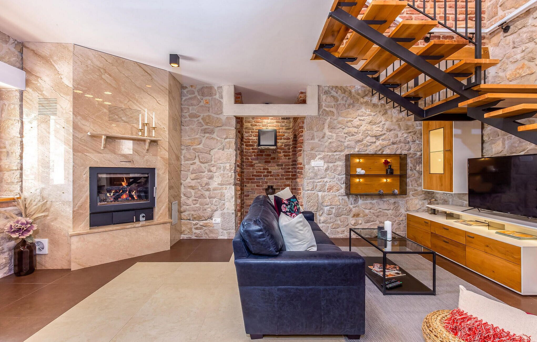 A modern living room with a stone fireplace, a black leather sofa, a glass coffee table, a wooden TV stand, and a wooden staircase.