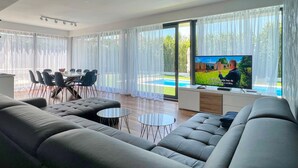 A modern living room with a large TV, grey sofas, a dining area with chairs, and a view of a swimming pool.