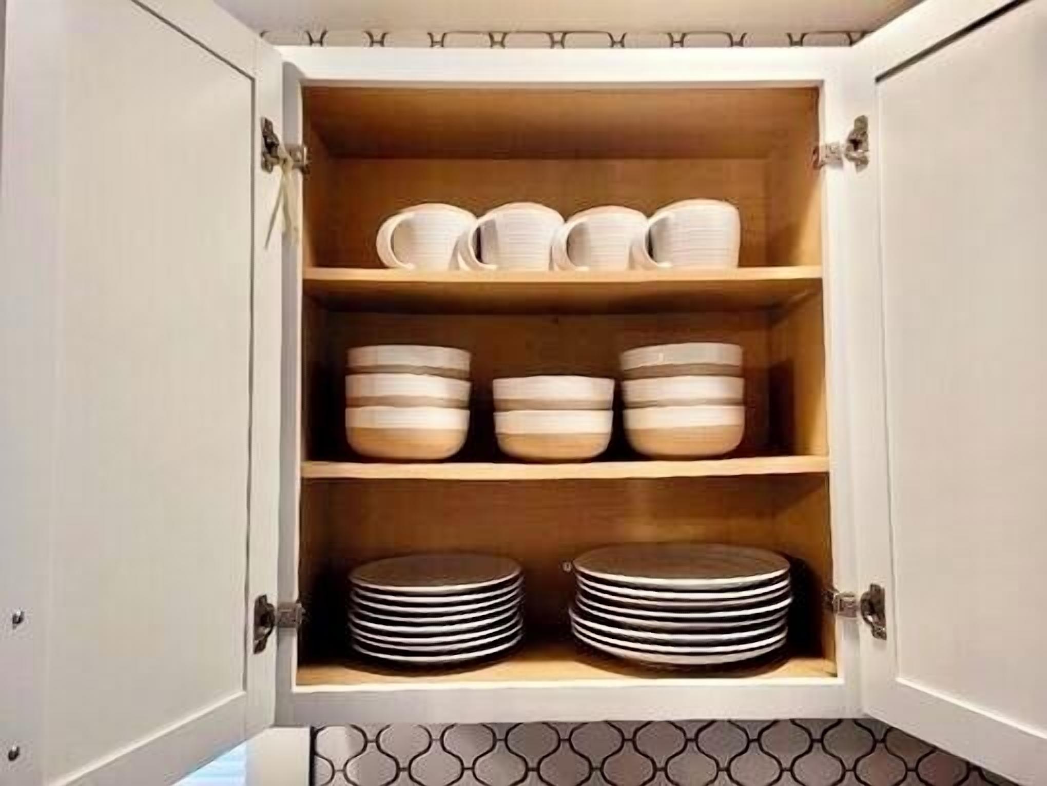 A kitchen cabinet with three shelves, each holding bowls, cups, and plates.