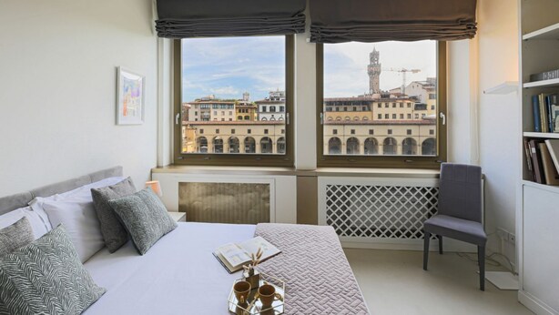 Un dormitorio con cama, una silla, un ventanal con vistas a edificios y un radiador.