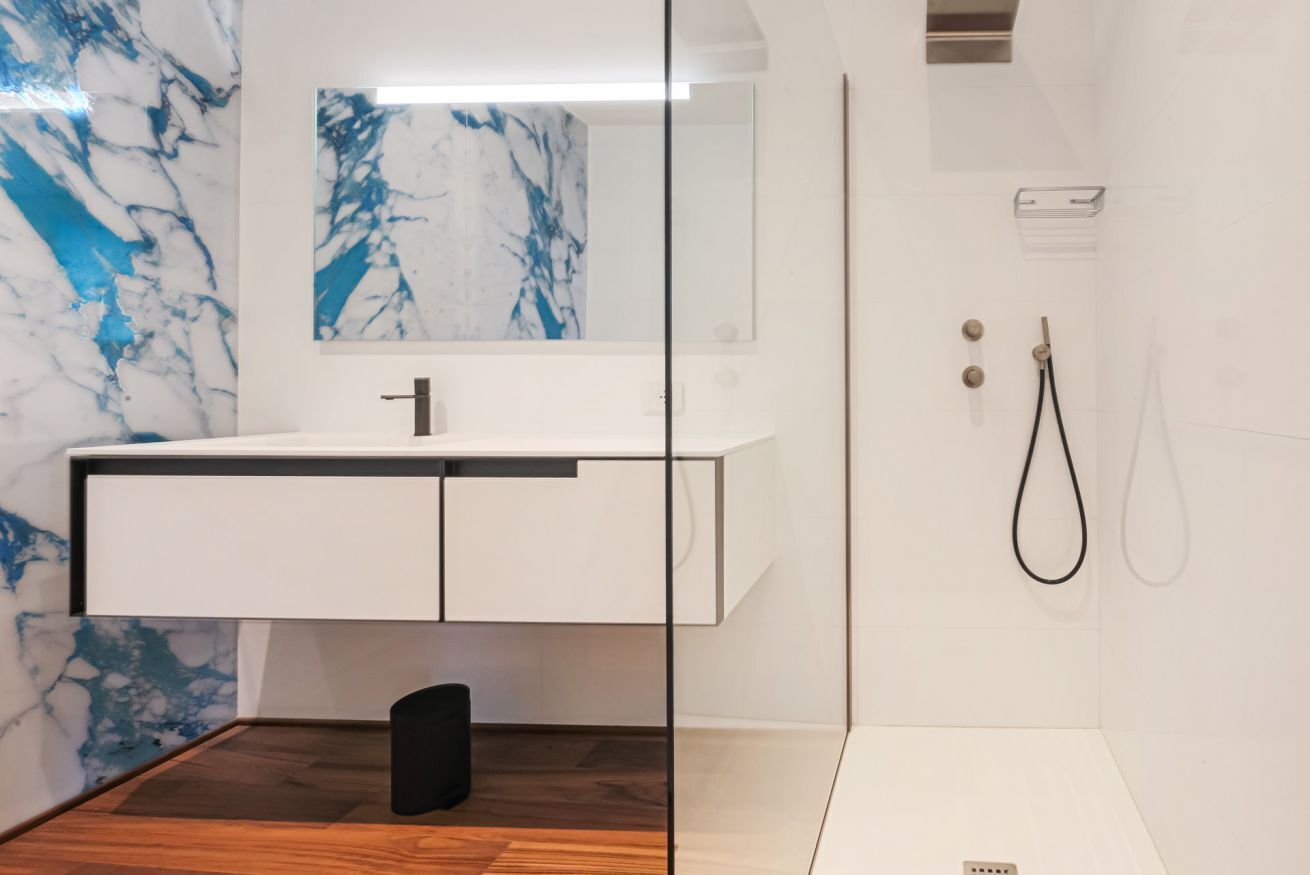 Un baño moderno con detalles de mármol, un tocador blanco, cabina de ducha de vidrio y suelo de madera.
