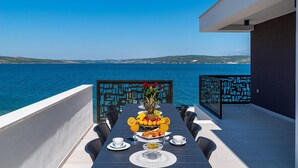 Et spisebord dækket med frugt, glas og tallerkener på en balkon med udsigt over havet.