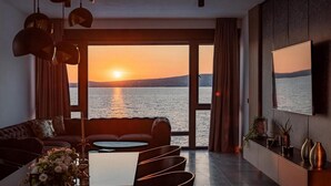 En moderne stue med sofa, sofabord og dekorative genstande. Et stort vindue giver udsigt til solnedgang over vandet.