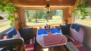 A cozy RV interior with a dining table, chairs, and a large window offering a view of a wooded area. - Boho Caravan themed, Vintage 1961 Oasis Camper (Monroe)