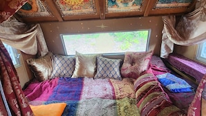 A cozy bed with a colorful quilt and patterned pillows, a window with a view of greenery, and a ceiling with decorative panels. - Boho Caravan themed, Vintage 1961 Oasis Camper (Monroe)
