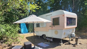 A vintage camper parked in a wooded area with a tent and a table with chairs set up nearby. - Boho Caravan themed, Vintage 1961 Oasis Camper (Monroe)