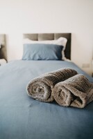 A neatly made bed with a blue bedspread and two rolled-up towels on top.