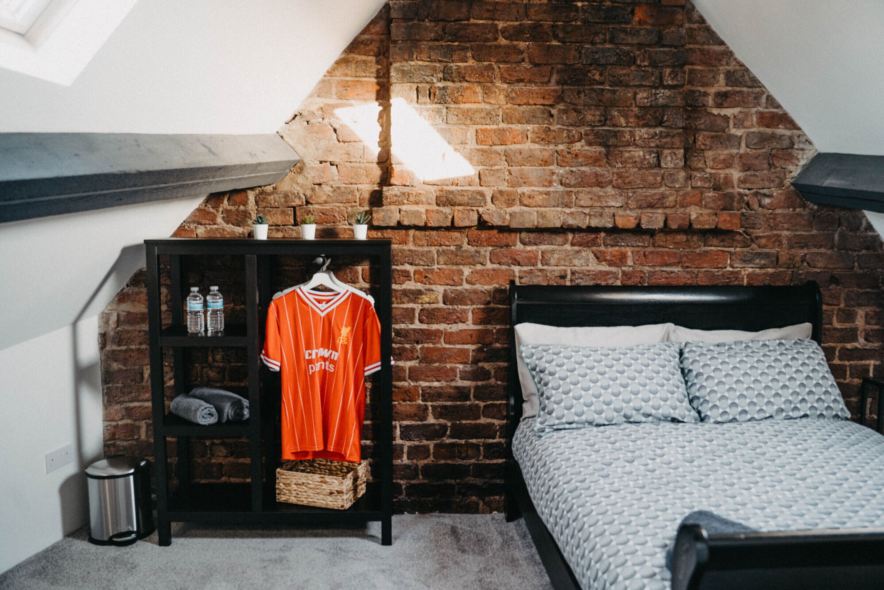 A bedroom with a brick wall, a bed with patterned bedding, a shelf with a jersey, and a small trash bin.