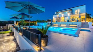 Ein modernes, zweistöckiges Haus mit Swimmingpool, Sonnenschirmen im Freien und einer gut beleuchteten Terrasse.