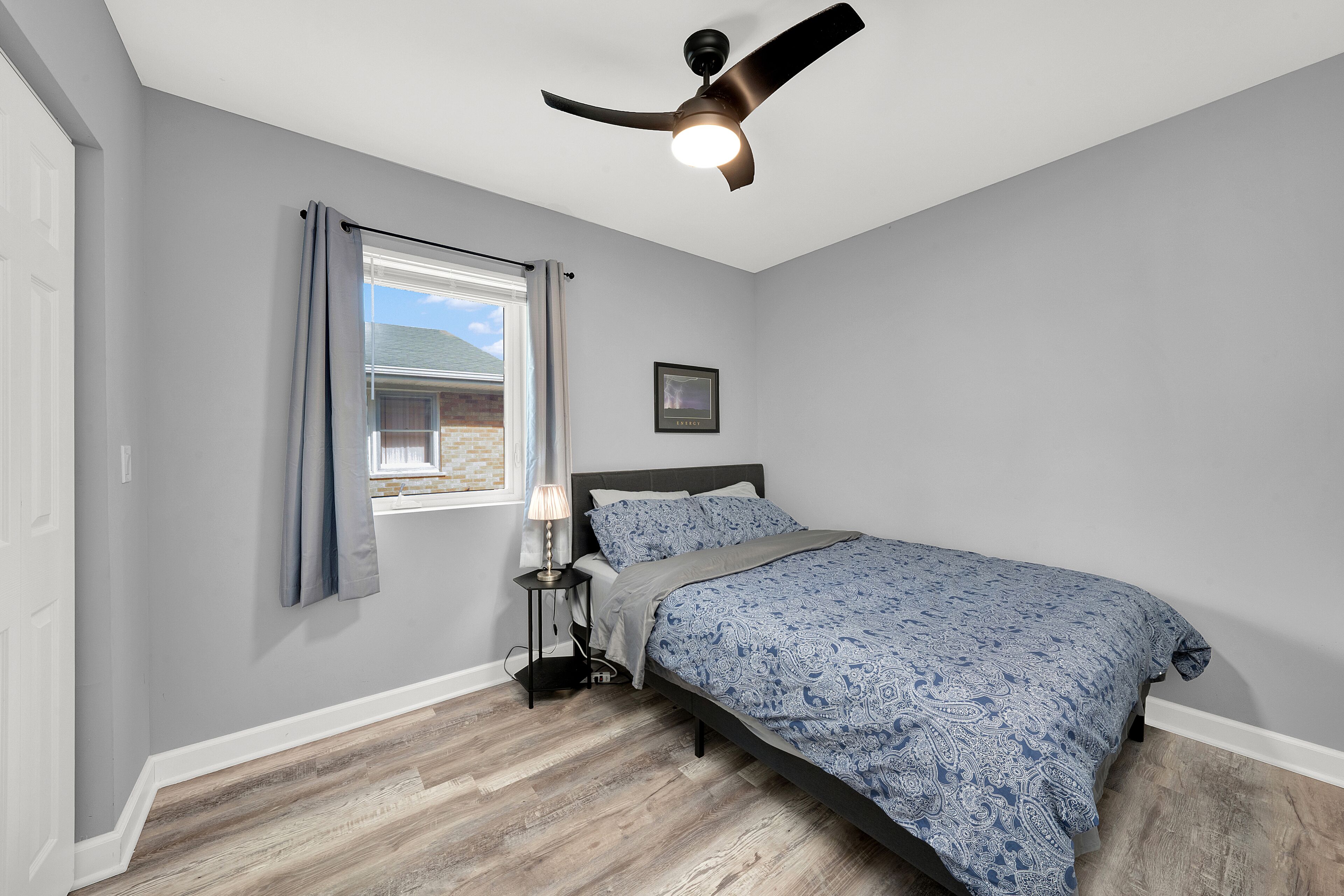 A bedroom with a bed, a ceiling fan, a window with curtains, and a small bedside table with a lamp.