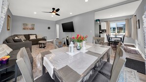 Ein modernes Wohnzimmer mit grauem Sofa, einem Esstisch mit Stühlen, einem Deckenventilator und einem an der Wand montierten Fernseher.