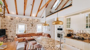A modern kitchen with a stone wall, wooden beams, a central island with bar stools, and white cabinetry. - Villa Mediterranean Estate (Uskoplje)