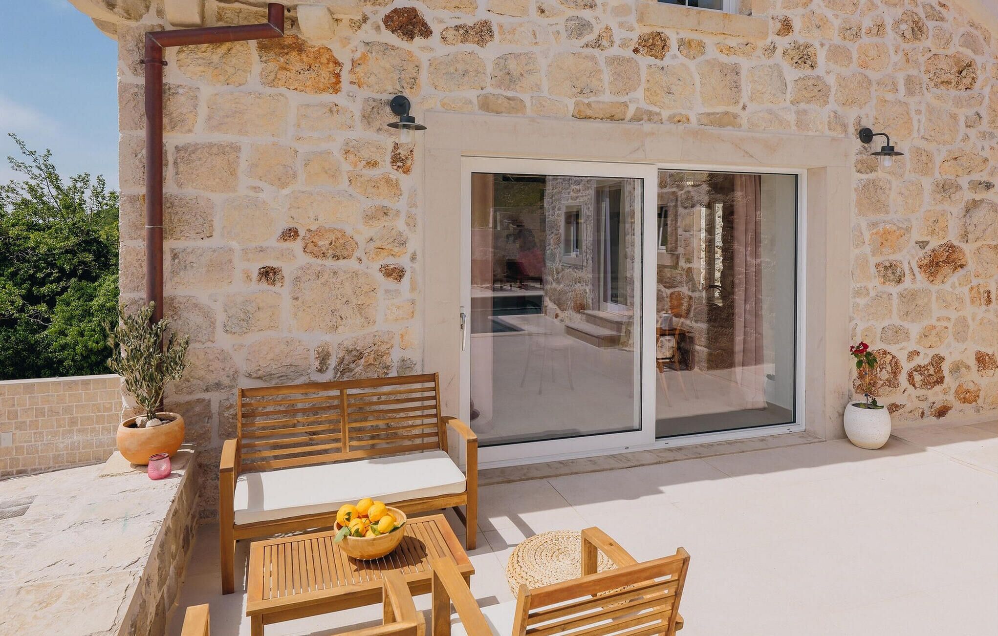 Un edificio de piedra con puerta de cristal que da a un patio. Hay bancos de madera y una mesa con un cuenco de fruta.
