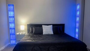 Una cama con una colcha oscura y dos almohadas, una mesita de noche con una lámpara, y una pared con paneles de iluminación azul verticales.
