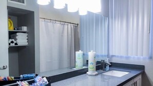 Un baño de hotel con un espejo grande, dos lavamanos y una cortina de ducha. Hay artículos de aseo en la encimera y una cesta con toallas.