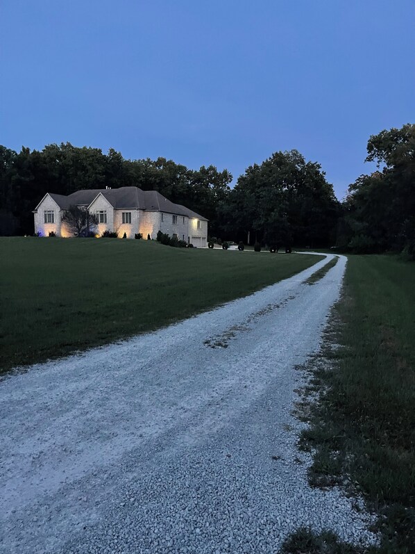 A gravel driveway leading to a house with a lawn and trees in the background.