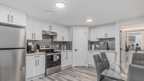 A modern kitchen with white cabinets, stainless steel appliances, and a glass dining table. - Kozy In Keswick (Suite) (Edmonton)