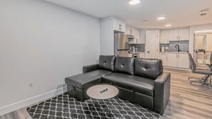 A modern living room with a sectional sofa, a small round coffee table, and a kitchen in the background. - Kozy In Keswick (Suite) (Edmonton)