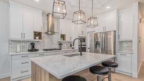 A modern kitchen with white cabinetry, a large island with bar stools, a stainless steel refrigerator, and hexagonal tile backsplash. - Kozy In Keswick (Upper Suite) (Edmonton)