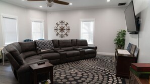 A living room with a sectional sofa, a coffee table, a rug with a polka dot pattern, a ceiling fan, and a flat-screen TV mounted on the wall.