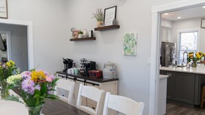 A modern kitchen with white cabinetry, a dining area with a wooden table and chairs, and a kitchen island with a coffee machine and snacks.
