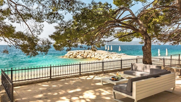 A coastal outdoor seating area with a stone floor, a white sofa, and a table with fruits and drinks.