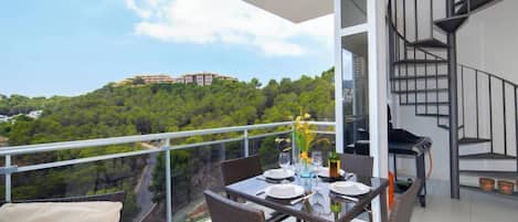 A balcony with a dining table set for four, overlooking a lush green hillside with buildings in the distance.