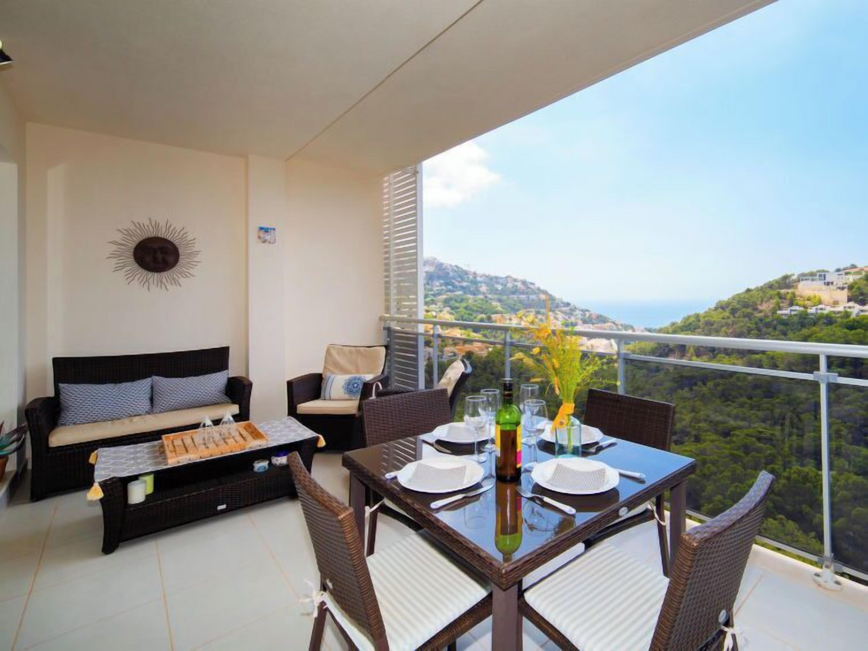 A balcony with a dining table set for two, a sofa, and a sun decoration on the wall.