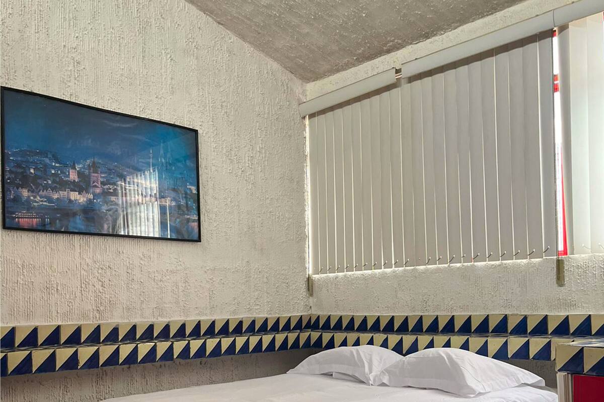 Quarto com uma cama, pintura de paisagem urbana, janela com persianas e parede estampada.