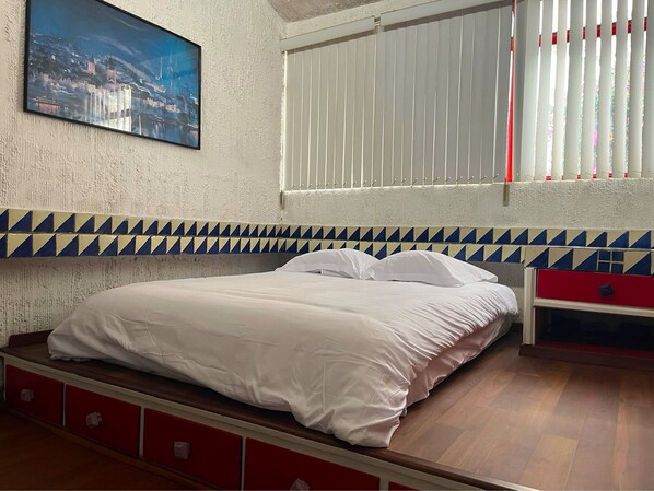 Une chambre à coucher avec un lit, une fenêtre avec des stores, une tête de lit rouge et bleue à motifs, et un tableau représentant un paysage urbain accroché au mur.