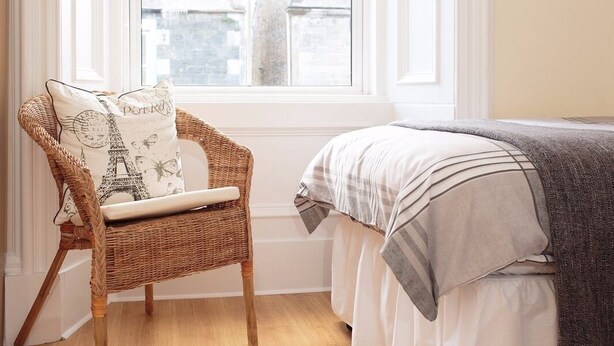 A wicker chair with a cushion featuring the Eiffel Tower, positioned next to a bed with a striped blanket. - The Corner House (Peebles)