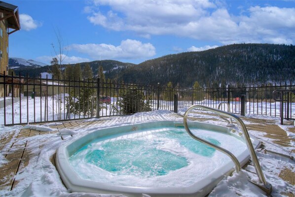 A hot tub surrounded by a wooden deck with snow on the ground, a metal fence, and a mountainous landscape in the background.