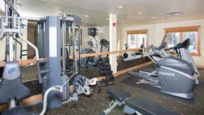Ein Fitnessstudio mit unterschiedlichen Trainingsgeräten, einem Spiegel und Fenstern mit Blick auf Bäume.