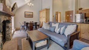Un salon chaleureux avec une cheminée en pierre, une table basse en bois, un canapé gris agrémenté de coussins et un coin repas avec des chaises et une table.