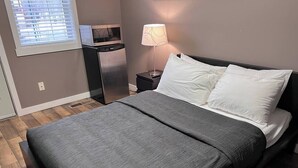 A modern bedroom with a bed, a nightstand, a lamp, and a small kitchenette. - Hotel Style 1 Bedroom & Bathroom (Fernie)