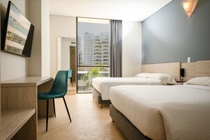 Habitación de hotel con escritorio, dos camas, una silla y vistas a edificios a través de un ventanal.